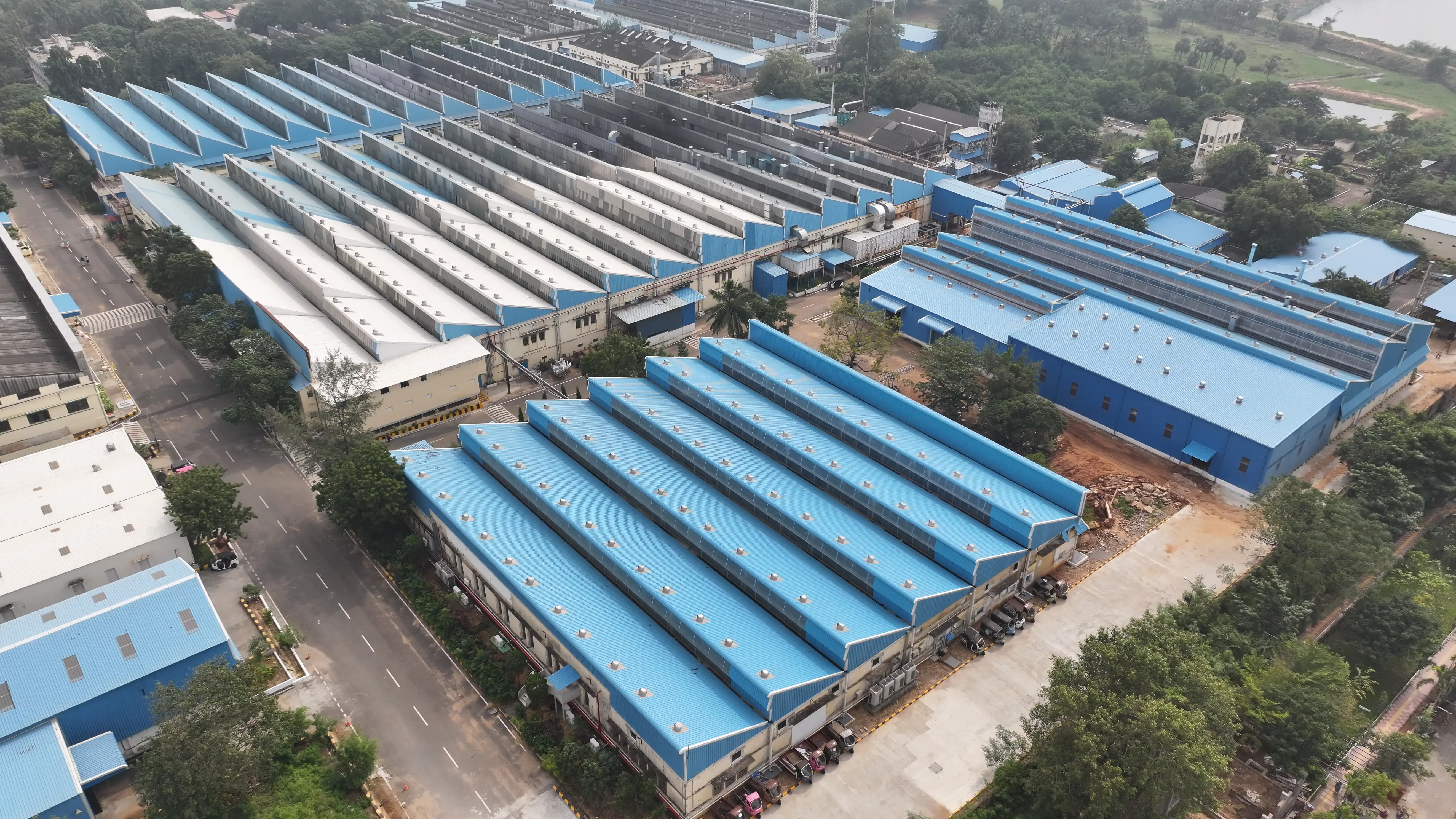 Aerial view of a large industrial complex with blue and white rooftops, surrounded by greenery and located near a waterbody.